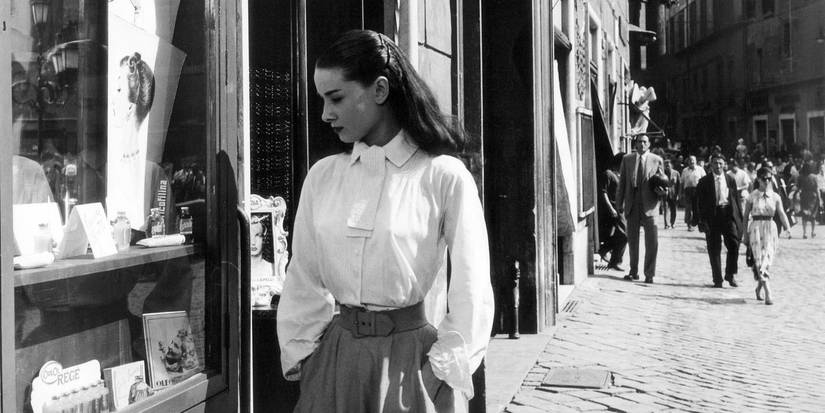 Princess Ann, played by Audrey Hepburn, wearing a long-sleeved white blouse, dark belt, and matching darker skirt, standing in the street looking through a shop window on her left in Roman Holiday