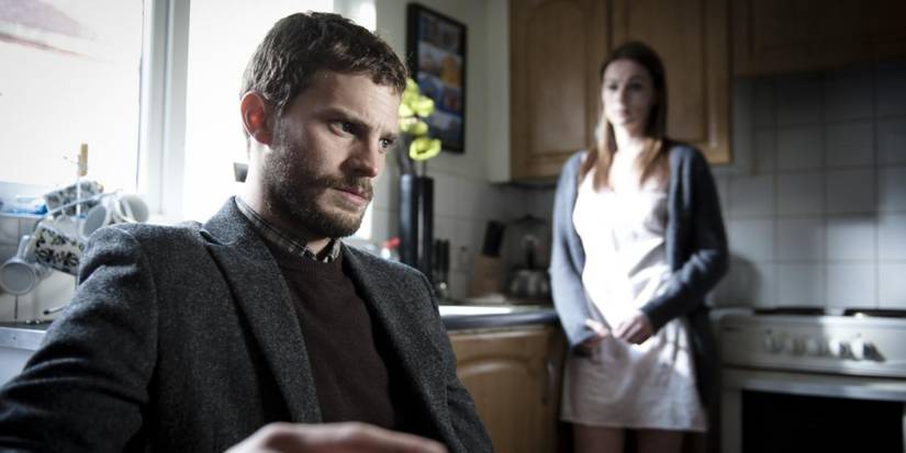 Jamie Dornan as Paul Spector sits in a kitchen with a woman standing behind him to his right in The Fall.