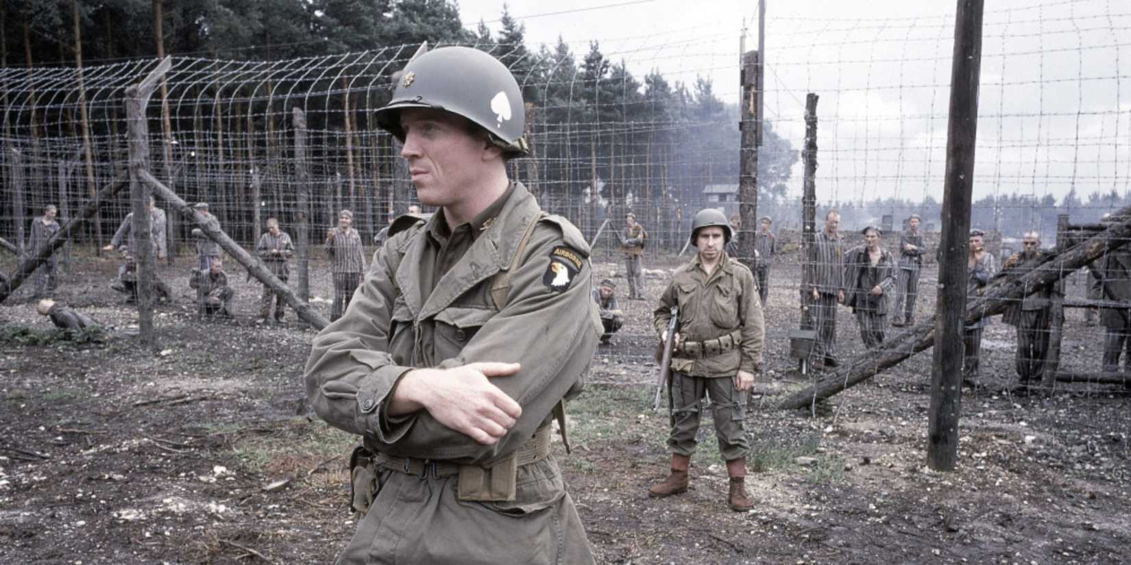 Damian Lewis as Major Richard Winters with arms folded in front of a tall wire fence in Band of Brothers.