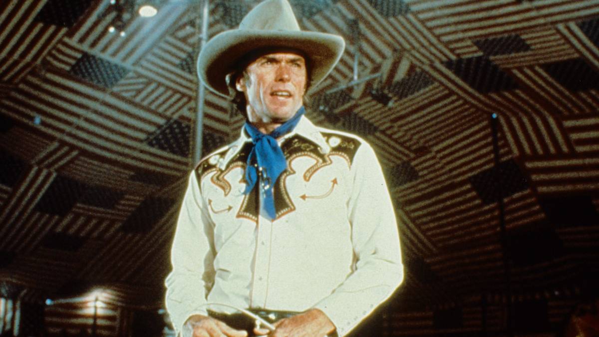 Bronco Billy stands in a Wild West show arena with the roof an overlapping mosaic of American flags.
