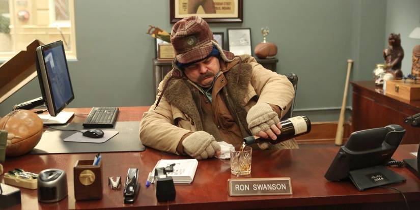 Nick Offerman as Ron Swanson in lots of clothes pours himself a drink at his desk in Parks and Recreation.