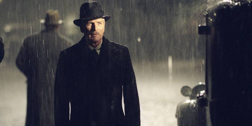 John Rooney (Paul Newman) stands wearing a hat and suit in the pouring rain as he stands by a car in 'Road to Perdition' (2002)
