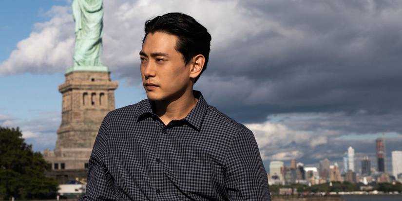 Teo Yoo as Hae Sung in Past Lives, standing in front of the Statue of Liberty.