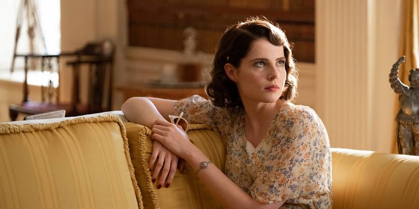 Frankie Derwent (Lucy Boynton) sitting on a couch in a flowery white dress and looking to the left with a dry expression in Why Didn't They Ask Evans?