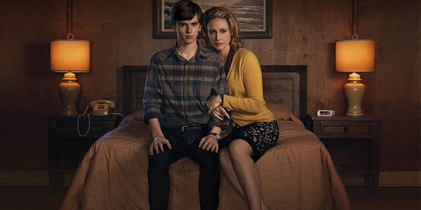Norman and Norma from 'Bates Motel', with arms linked, sit on a bed and look at the camera dramatically.