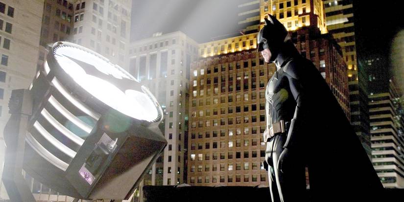 Christian Bale as Batman standing before the Bat-Signal at night in Batman Begins. 