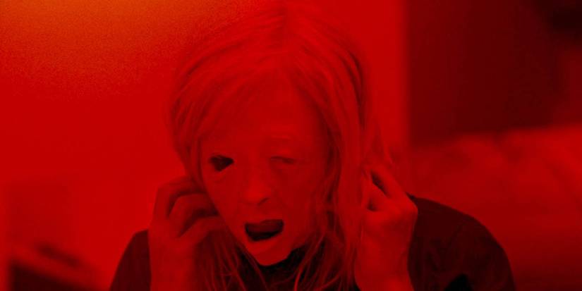 Andrea Riseborough tears a distorted mask off her face while cast in stark, red lighting in Possessor.