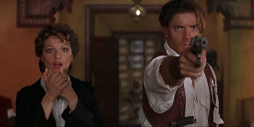 Rick O'Connell (Brendan Fraser) aiming a gun ahead with Evie Carnahan (Rachel Weisz) standing next to him with her hands near her gasping mouth, staring at the same offscreen thing he is, in The Mummy