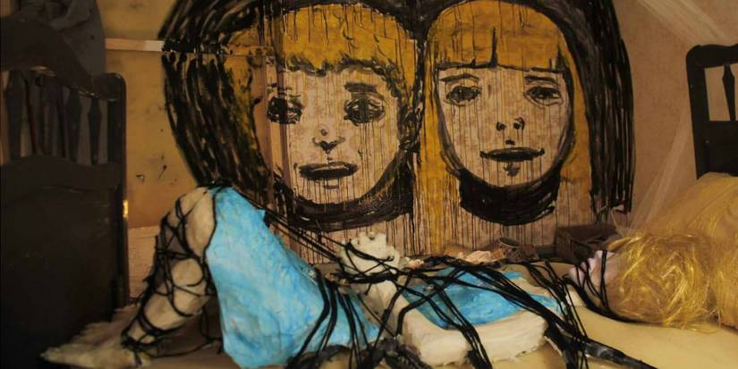 A young girl tied to the bed next to a wall with children's faces painted in The Wolf House-1