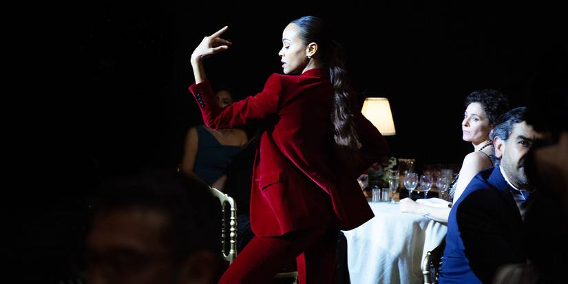 Zoë Saldaña dances in a red suit in a dark room around tables of people in Jacques Audiard's Emilia Pérez.