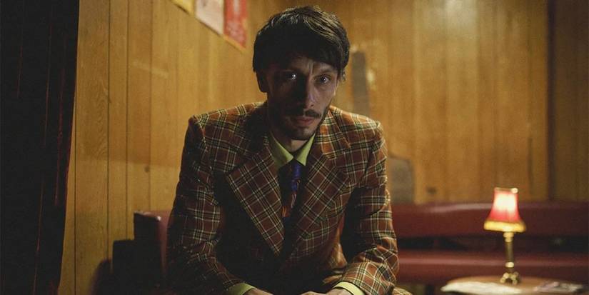 Donny sitting in a plaid suit with his arms resting on his legs in Baby Reindeer.