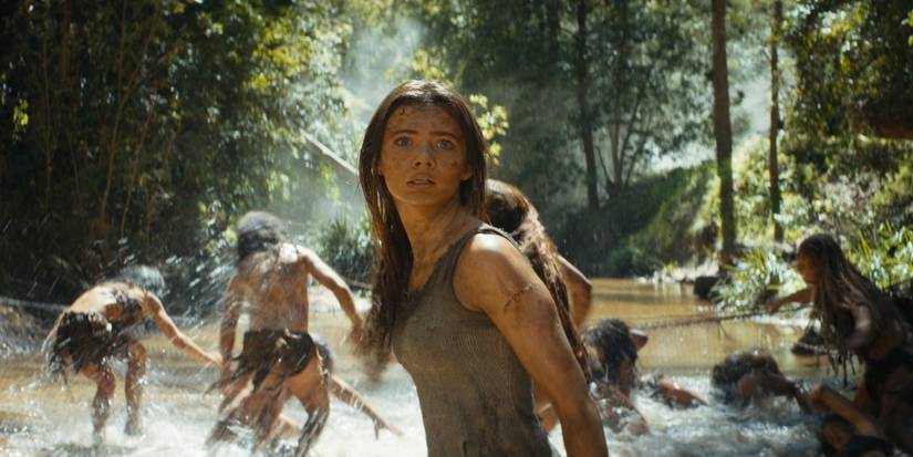Freya Allan as Mae standing in a dirty river while people run away behind her in Kingdom of the Planet of Apes