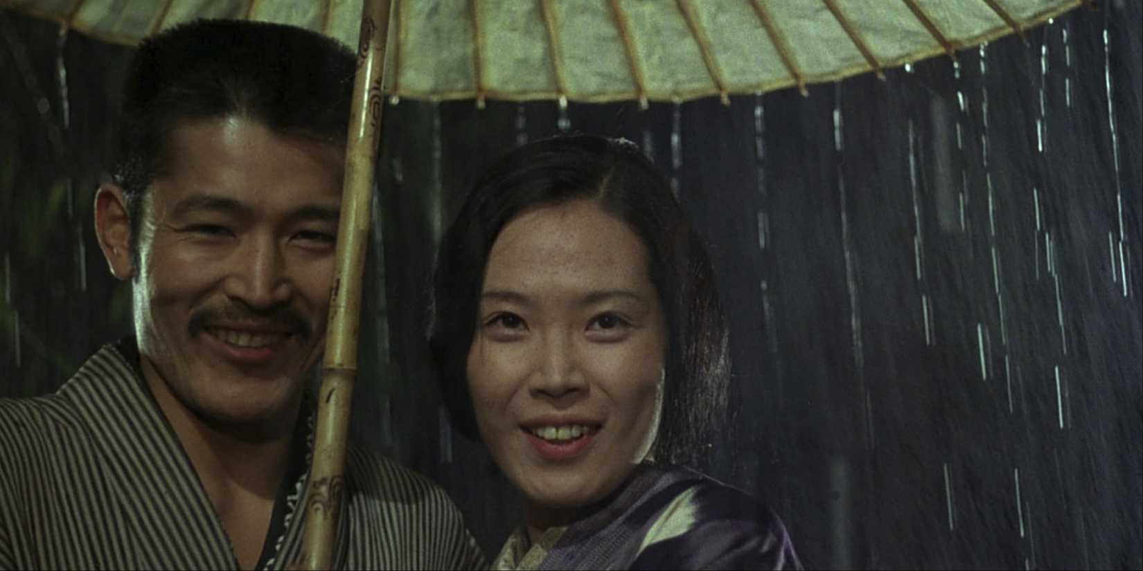 A man and woman stand under an umbrella looking toward the camera in In the Realm of the Senses