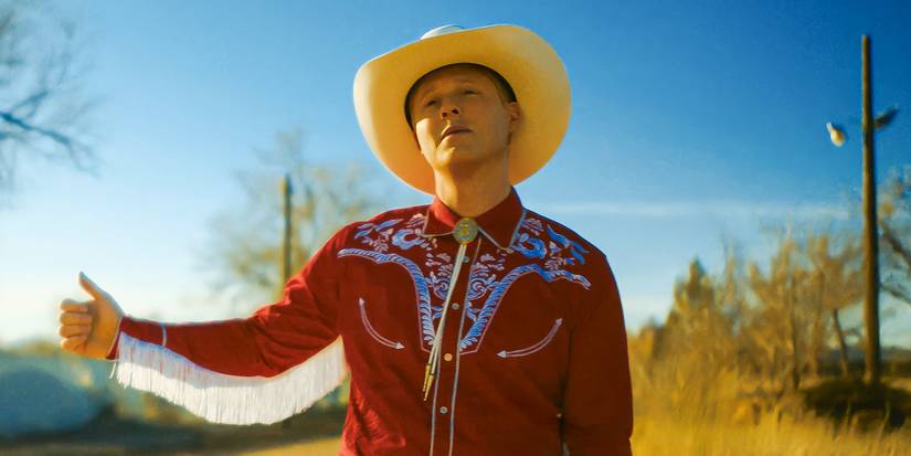 Noah Reid as Billy Tillerson wearing a red jacket and hitchhiking in Outer Range Season 2. 