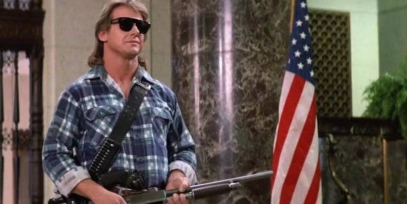 Roddy Piper with a shotgun wearing sunglasses standing next to an American flag in They Live.
