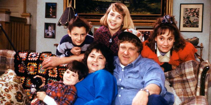 The cast of Roseanne sitting on a couch in the living room posing.