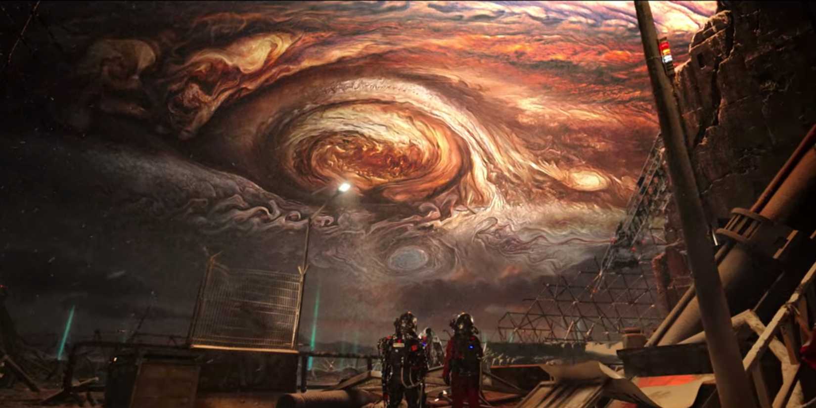 Two tiny figures on a destroyed earth surface look up at Jupiter, which fills the sky.