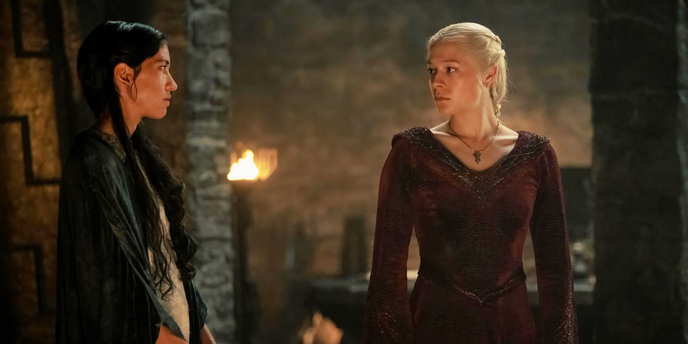 A woman (Mysaria) with black hair and a green silk dress talks to the woman to her right (Rhaenyra) who has light blonde hair and a red dress. The woman on the right looks stern, and they are standing in a stone room with a small fire lit in the background.