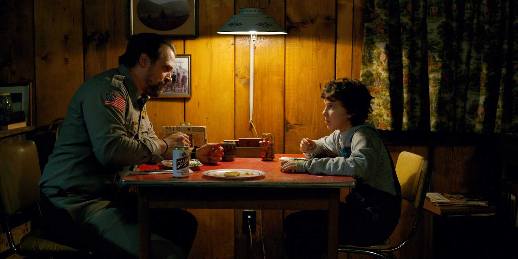 Hopper dan Eleven duduk di meja dan saling berhadapan dalam trik episode Stranger Things atau memperlakukan orang aneh.