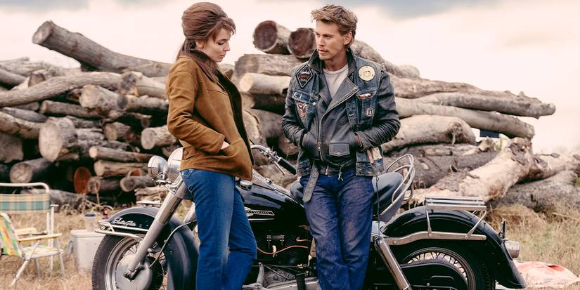 Jodie Comer stands near Austin Butler in front of a motorcycle and logs, both with hands in their pockets
