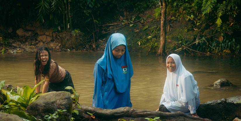Two girls wearing hijabs and one in a black skirt sitting by a river in Tiger Stripes