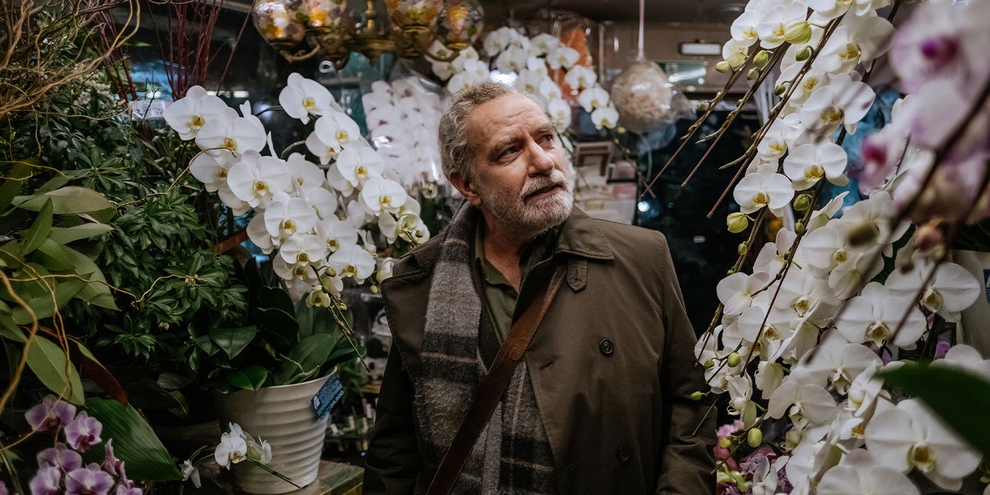A still of Egill Ólafsson in a coat walking down an aisle of white orchids smiling