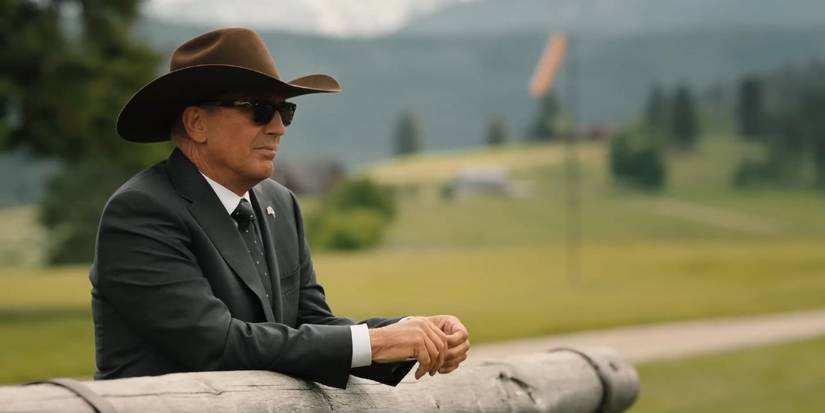 John Dutton (Kevin Costner) wearing sunglasses and a cowboy hat, leaning on a wood fence in Yellowstone.