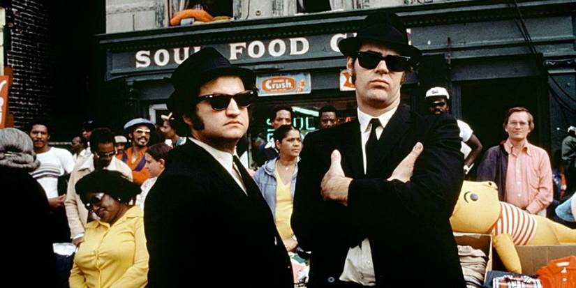 John Belushi and Dan Aykroyd in suits, hats, and sunglasses with people behind them in the Blues Brothers.