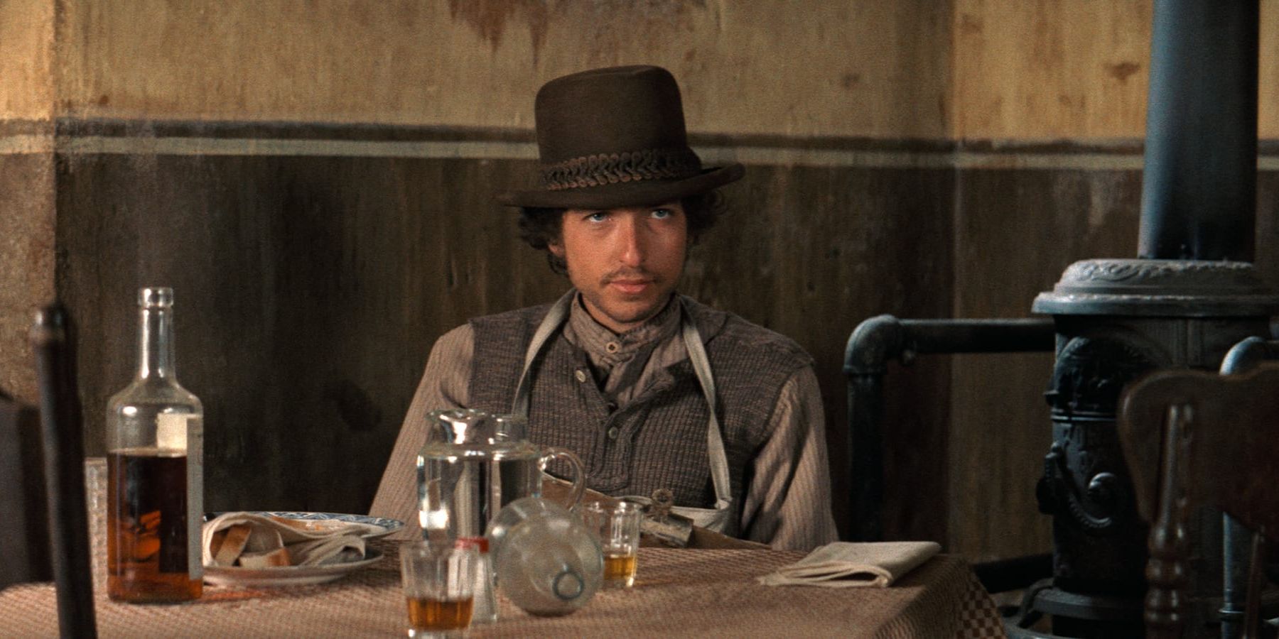 Alias (Bob Dylan) sits at a bar table surrounded by liquor bottles and glasses in 'Pat Garrett and Billy the Kid'