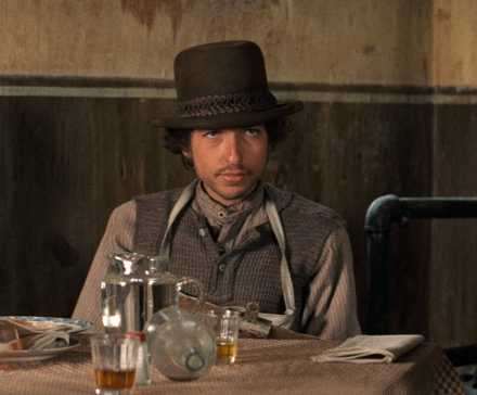 Alias (Bob Dylan) sits at a bar table surrounded by liquor bottles and glasses in 'Pat Garrett and Billy the Kid'
