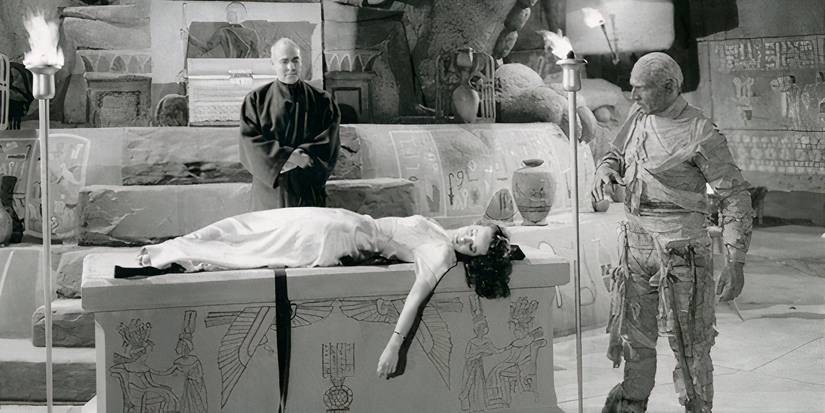 A man and a mummy holding a torch overlook a woman lying on a plinth 