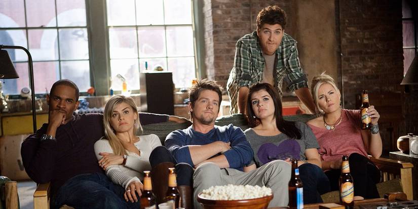 The cast sit on a couch and watch something with a bowl of popcorn and beer in Happy Endings.