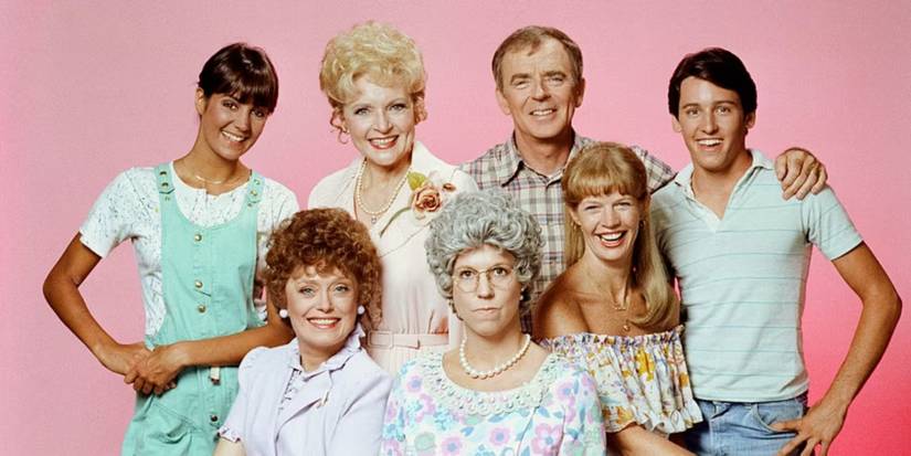 The cast of Mama's Family all smile except for Mama, as they look at the camera with a plain pink background.