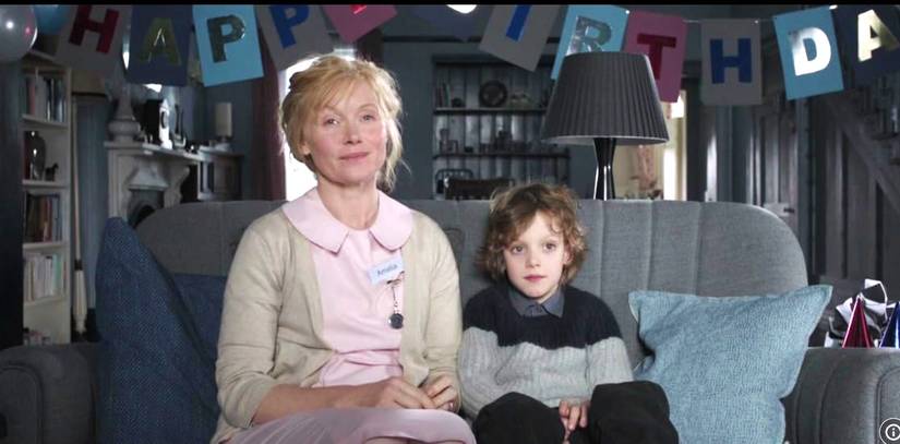 Essie Davis and Noah Wiseman as Amelia and Sam sitting together on a couch in front of a birthday banner in "The Babadook"