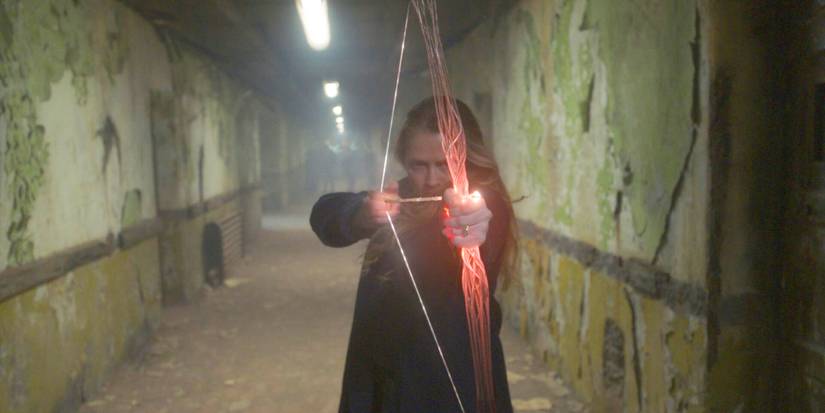 Diana Bishop (Teresa Palmer) aiming a red-tinted magical arrow down a long corridor of a decaying hospital in A Discovery of Witches Season 3