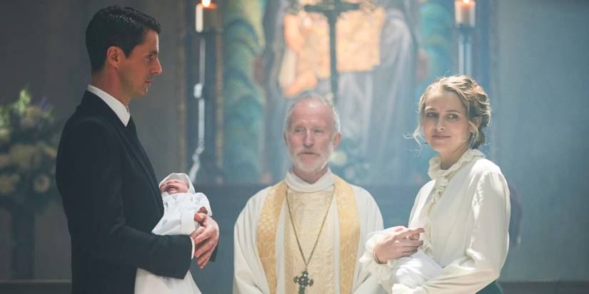 Matthew Clairmont (Matthew Goode) and Diana Bishop (Teresa Palmer) standing in front of a Catholic priest at their twins' christening ceremony, with Matthew holding one twin and standing on the left in a black suit and Diana holding the other baby and standing on the right in a white blouse, in A Discovery of Witches Season 3