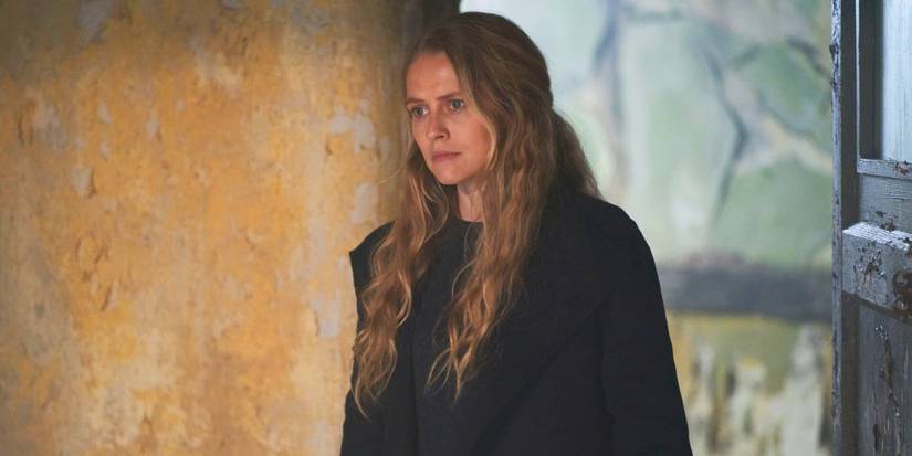 Diana Bishop (Teresa Palmer) standing in an old hospital room and looking concerned while wearing a dark coat and her long blonde hair down in A Discovery of Witches Season 3