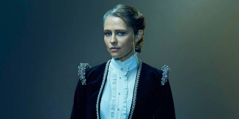 Diana Bishop (Teresa Palmer) facing the camera, standing against a blue-green tinted background and wearing a dark navy jacket over a white blouse with her blonde hair up, in A Discovery of Witches Season 3