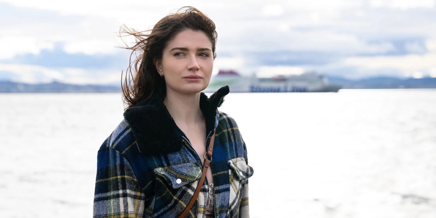 In a scene from Bad Sisters, Becca Garvey (Eve Hewson) stands in front of the ocean wearing a plaid coat and her hair blowing in the wind.