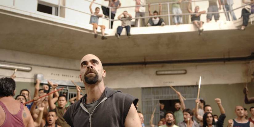 A man stands in a multi-tiered venue with people holding sticks in the background in Cell 211.