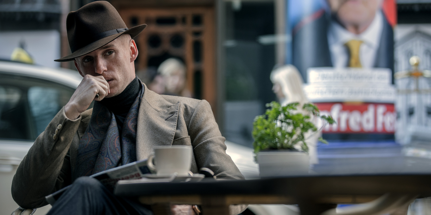 The Jackal sits in disguise at an outdoor cafe looking pensive in The Day of the Jackal.