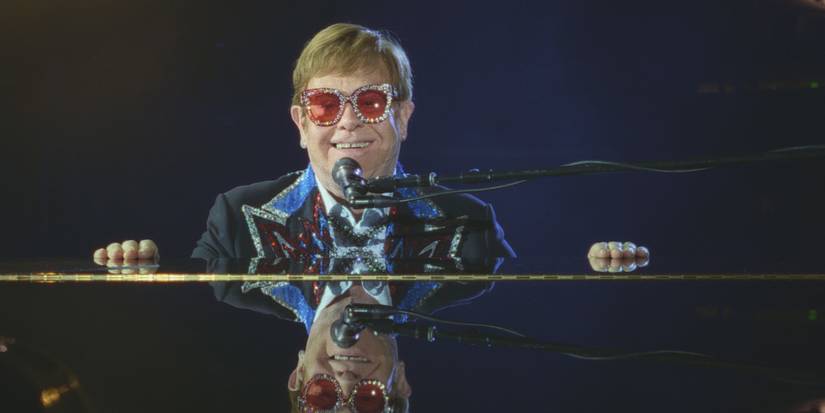 Elton John smiling in his bejeweled glasses at a grand piano on stage