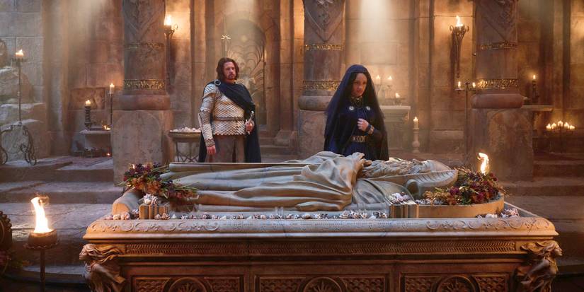 A aide shot of Miriel (Cynthia Addai-Robinson) standing at her father's tomb in a dark mourning outfit with Elendil (Lloyd Owen) standing several steps behind her and to her left in his white armor and cloak in Rings of Power Season 2.