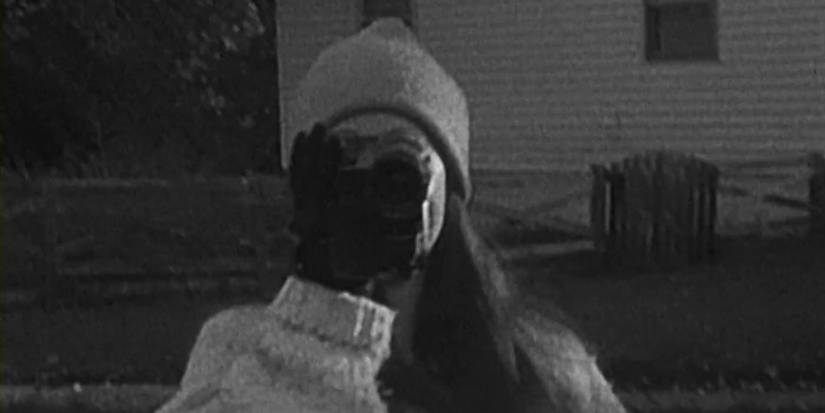A black-and-white image of Heather from 'The Blair Witch Project' holding a video camera in front of her face.