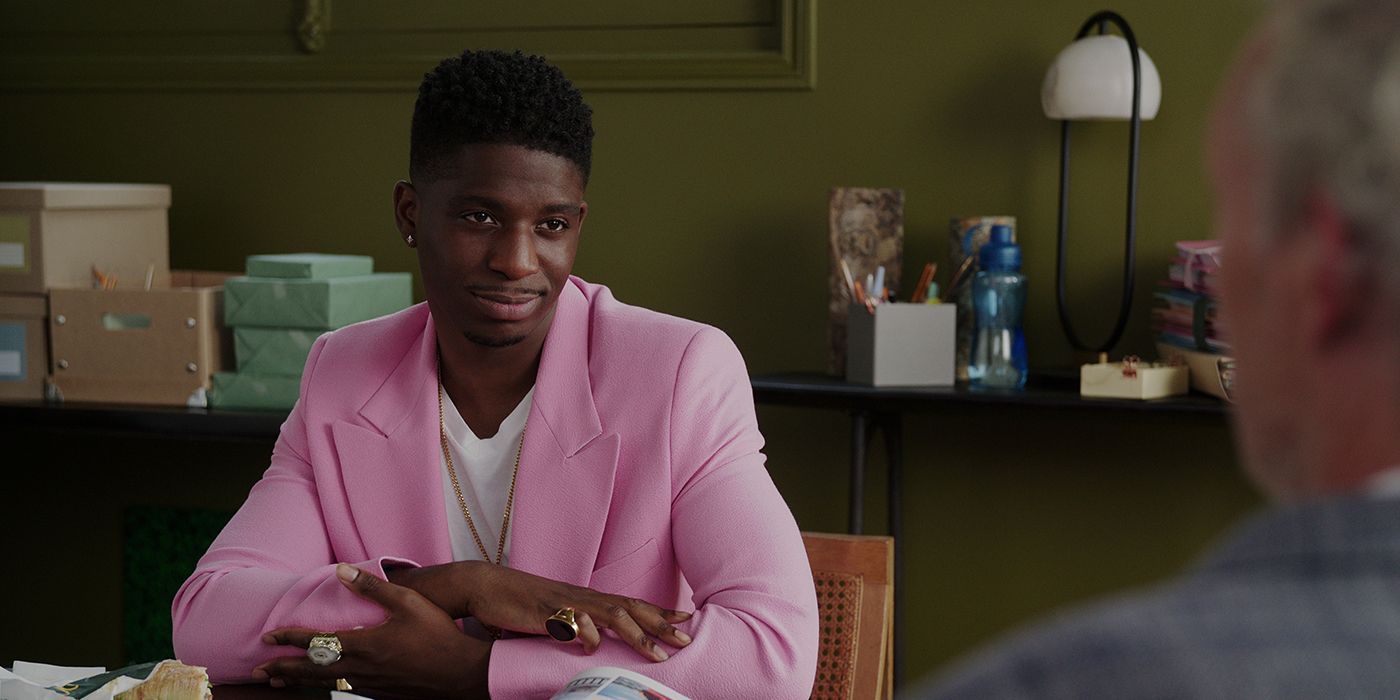 Julien at his desk, hands folded in front wearing a pink blazer on Emily in Paris