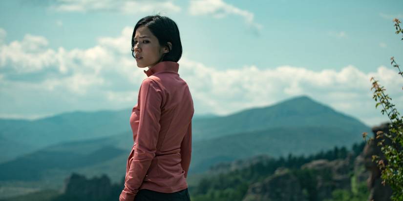 Kelsey Asbille as Iris, wearing a pink sweater, standing on the edge of a cliff, and looking behind her in Netflix's Don't Move 