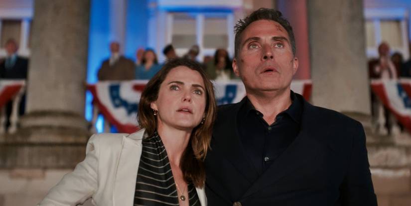 Keri Russell dan Rufus Sewell di luar gedung bersama orang-orang, melihat ke arah The Diplomat Musim 2.