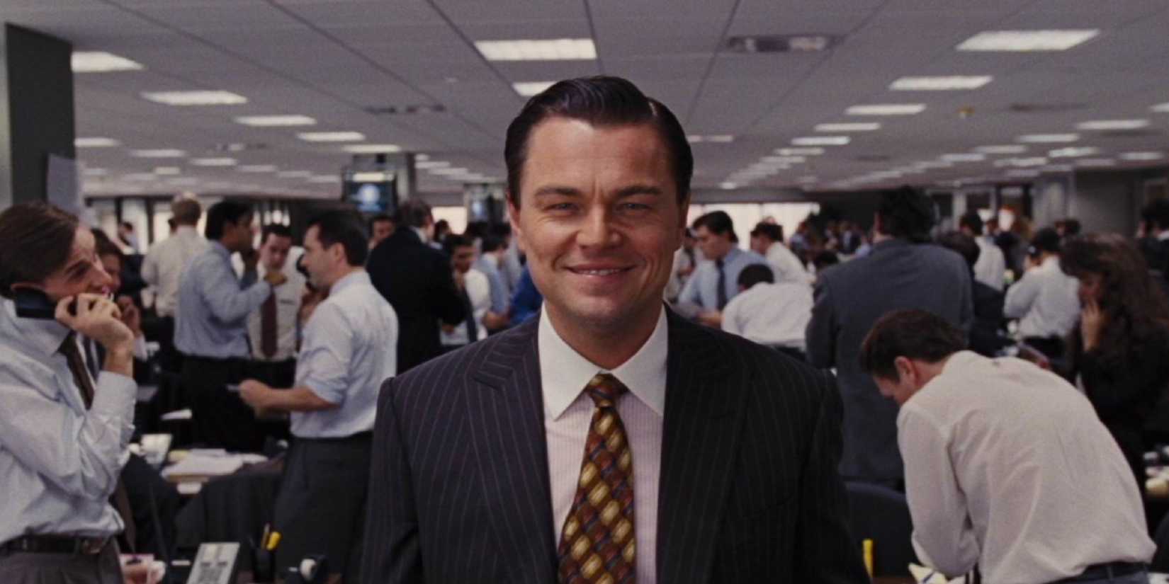 Leonardo DiCaprio as Jordan Belfort smiling into the camera in a busy office in Wolf of Wall Street.