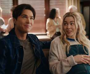 Denny sits next to a pregnant Lizzy at a table in a venue and smiles in Virgin River season 6.