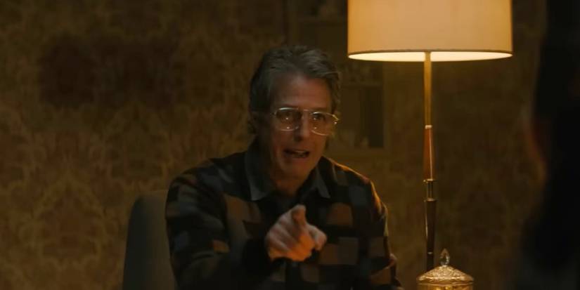 Hugh Grant as Mr. Reed pointing a finger towards the camera while sitting next to a lamp giving off yellow light in 'Heretic'